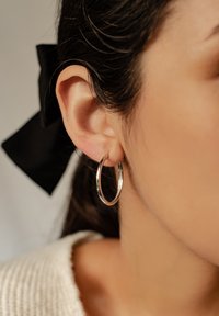 Pendiente de aro plateado colgando de una oreja, con una superficie lisa y reflectante. Se puede ver un lazo negro en el cabello detrás de la oreja.