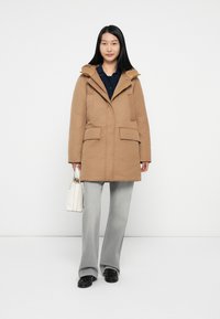 Bruine gewatteerde parka met een ritssluiting en drukknoopsluiting, twee voorzakken en een licht gestructureerde pasvorm, gecombineerd met lichtgrijze broek en een witte tas.