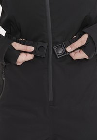 Svart jumpsuit med dragkedja fram och justerbar midja med en platt svart spänne. Texturerat tyg med åtsittande ärmslut. Minimalistisk design.