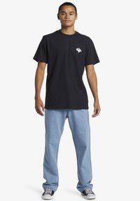 Crna pamučna majica s malim bijelim grafičkim uzorkom na prsima, u kombinaciji s svijetloplavim trapericama i crno-bijelim tenisicama.