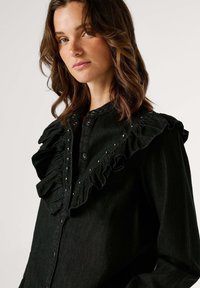 Čierna denimová košeľa s nariaseným golierom ozdobeným striebornými cvočkami, zapínaním na gombíky a mäkkým materiálom. Strih je prispôsobený a priliehavý.