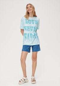 Lichtblauwe tie-dye T-shirt met gedurfde turquoise tekst "LOVE NEVER FADES", gedragen met denim shorts en chunky witte sandalen.