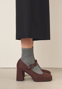 Bruna högklackade skor i mocka med rundad tå och spänne, använda över grå knästrumpor, kombinerade med en svart kjol.