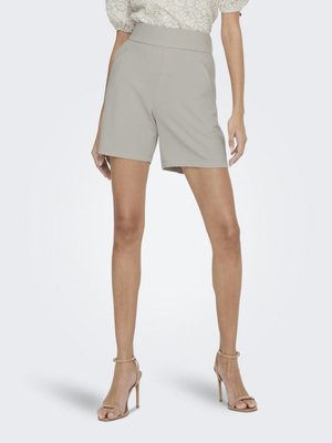 Vrouw draagt een beige short met hoge taille, een witte blouse met structuur en beige enkellaarsjes met hoge hak, staand tegen een effen witte achtergrond.