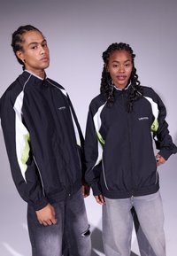 Casacos à prova de vento pretos com detalhes em branco e verde lima, fecho éclair à frente e punhos elásticos. Os jeans de ganga do modelo apresentam um aspeto desgastado.