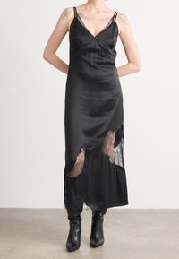 Femme portant une robe nuisette en satin noire avec des panneaux en dentelle et des bottes noires à talons, debout avec les mains derrière le dos sur un fond uni.