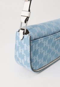 Bolso de hombro de denim azul con un patrón de pata de gallo, que presenta una correa blanca ajustable y herrajes plateados. Textura suave y forma rectangular.
