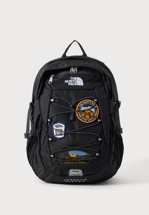 Schwarzer North Face-Rucksack mit vorderem Spanngurt, drei bunten Aufnähern und einem oberen Griff vor neutralem Hintergrund.