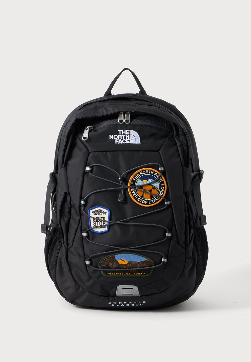 Μαύρο σακίδιο North Face με μπροστινό λάστιχο, τρία πολύχρωμα patches και επάνω χερούλι πάνω σε μονόχρωμο φόντο.