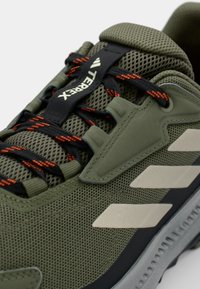 Olivgrüner Sportschuh mit Mesh-Obermaterial, schwarzen und orangenen Schnürsenkeln, hellgrauen Akzenten und Gummisohlendetails. Auffällige TERREX-Branding.