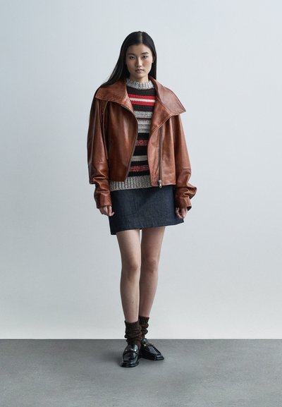 Brązowa skórzana krótka kurtka z dużym kołnierzem, noszona na paskowanym dzianinowym swetrze, zestawiona z ciemną spódnicą dżinsową i czarnymi butami.