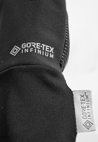 Sort stof med en glat tekstur, der fremhæver "GORE-TEX INFINIUM" logoet i grå. Et lille gråt mærke viser også det samme branding.