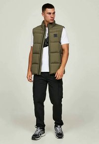 Jonge man in een olijfgroene gewatteerde vest over een witte graphic t-shirt, zwarte cargobroek en zwart-witte sneakers, staand en naar links kijkend.