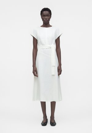 Žena stojaca čelom vpred má na sebe biele midi šaty s krátkym rukávom a opaskom v páse, v kombinácii s čiernymi plochými topánkami.