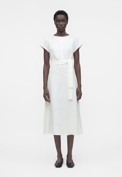 Femme debout, face à l’avant, portant une robe midi blanche à manches courtes avec une ceinture à la taille, associée à des chaussures plates noires.