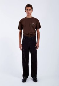 Maglietta di cotone marrone a maniche corte; pantaloni in velluto a coste neri con taglio dritto; scarpe nere; design minimalista; sfondo uniforme.