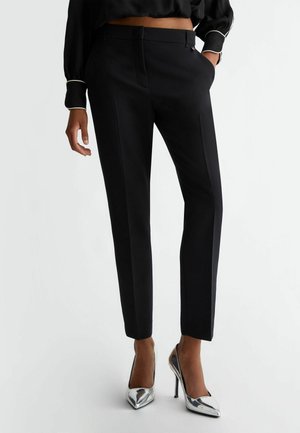 Femme portant un pantalon noir court et cintré, un chemisier noir avec une bordure blanche et des chaussures argentées à talons hauts et bouts pointus.