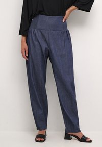 Tummat siniset denimgatsoitushousut, joissa on korkea smokkivyötärö, löysä istuvuus ja kapenevat lahkeet. Yhdistetty mustiin korkokenkiin.