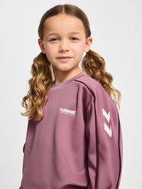 Młoda dziewczyna z długimi falowanymi włosami splecionymi w kucyki, ubrana w mauve bluzę Hummel Performance z białym logo w kształcie szewrona na rękawie.