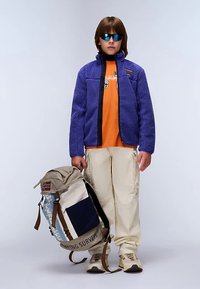 Giovane che indossa una giacca in pile blu, una maglietta arancione, pantaloni cargo beige, scarpe da ginnastica e occhiali da sole, mentre tiene uno zaino grande beige e blu.