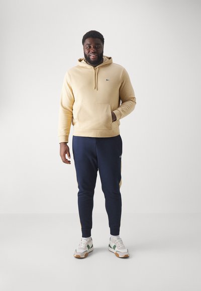 Hoodie beige avec une poche avant et un logo, associé à un jogging bleu marine avec des accents jaunes sur les côtés. Baskets blanches avec des détails verts.