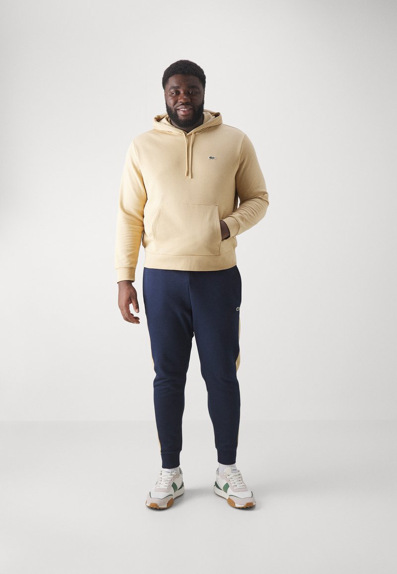 Beige Kapuzenpullover mit einer Vordertasche und einem Logo, kombiniert mit marineblauen Jogginghosen mit gelben seitlichen Akzenten. Weiße Sneakers mit grünen Details.