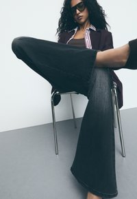 Široke sivo-denimske hlače z neobrobnim robom, v kombinaciji z burgundskim blazerjem in črtasto srajco. Model sedi na kromirani stolu.