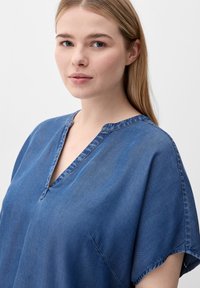 Blue denim top with a V-neckline, short sleeves, and a relaxed fit. The fabric has a smooth texture and subtle sheen, with contrast stitching.