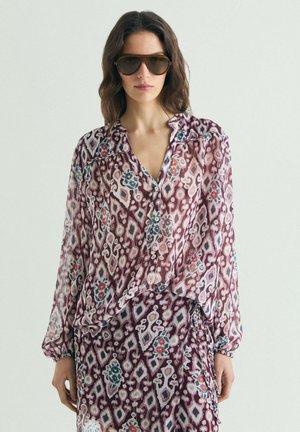 Mujer con cabello castaño ondulado, que lleva grandes gafas de sol marrones y un conjunto a juego de blusa y falda con estampado en tonos burdeos, rosa y azul.