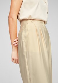 Beige veckade byxor gjorda av ett slätt tyg, med en skräddarsydd passform och hög midja. Tyget verkar lätt och flytande.