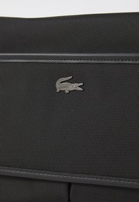 Borsa in tessuto nero con texture, caratterizzata da un logo in metallo a forma di coccodrillo, design elegante con piping grigio e un'estetica minimalista.