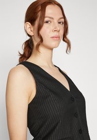 Melna bezpiedurkņu veste ar V veida kakla izgriezumu, svītrainu rakstu un trim melnām pogām priekšpusē. Gluda tekstūra ar izteiktām plecu līnijām.