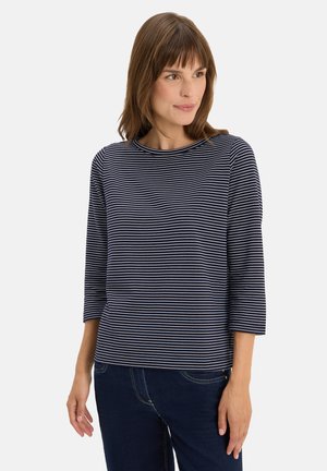 Femme portant un haut à rayures bleu marine et blanches à manches trois-quarts et un jean bleu foncé, se tenant contre un fond blanc uni.