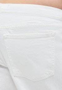 Κοντινή λήψη της πίσω τσέπης και της ζώνης μιας λευκής τζιν παντελόνας σε ανοιχτόχρωμο δέρμα, που δείχνει την υφή του υφάσματος και τις λεπτομέρειες στη ραφή.