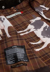 Καφέ καρό ύφασμα με εικονογραφήσεις αγελάδων, με μαύρη ετικέτα φροντίδας συνημμένη. Περιλαμβάνει γιακά από χοντρή ύφανση και πολύχρωμες λεπτομέρειες ετικετών.