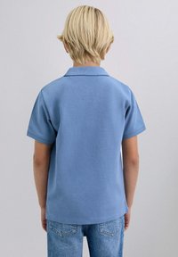 Lyseblå polo-skjorte med korte ærmer, krave og struktureret stof, parret med lyseblå denim jeans. Kun bagside.
