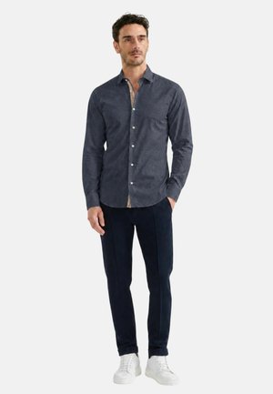 Uomo con camicia blu scuro abbottonata, pantaloni scuri e sneaker bianche, in piedi con una mano in tasca su uno sfondo semplice.