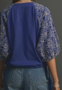 Modrý top s texturovanými nabíranými rukávy zdobenými propracovanou světlou výšivkou; látka působí jemně, s nastavitelnými šňůrkami na spodním okraji.