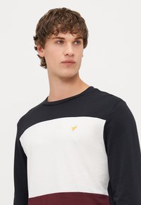 Langärmliges Hemd mit horizontalen Streifen in Schwarz, Weiß und Bordeaux. Aus Baumwollmaterial mit kleinem gelben gesticktem Logo auf der Brust.