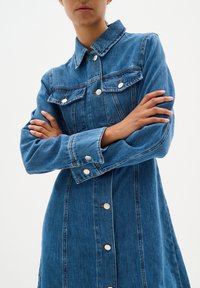 Robe en denim avec un col, des manches longues et des poches avant, présentant un délavage léger, des accents de boutons en argent et une silhouette ajustée.