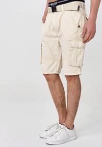 Beige Cargo-Shorts mit mehreren Taschen, genähten Details und einer regulären Passform, kombiniert mit weißen Sneakers und einem dunklen Bund.