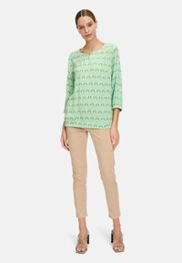 Blusa verde a fantasia con maniche a tre quarti; abbinata a pantaloni beige aderenti e sandali con tacco a strappo. Tessuto dalla superficie liscia.