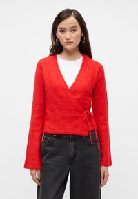 Červený svetr s omotáváním, vyrobený z jemného, texturovaného pletení, s dlouhými rukávy a detailem na zavázání v pase, spárovaný s černými džíny s vysokým pasem.