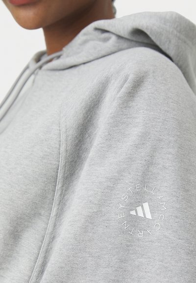 Primer plano de una persona que lleva una sudadera con capucha gris claro con un logo circular en la manga que presenta franjas estilizadas y texto.