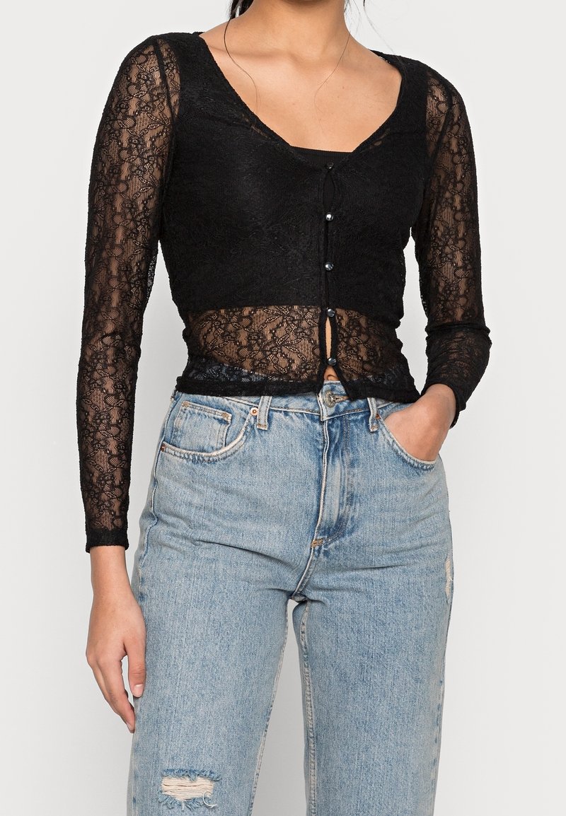 Top in pizzo nero a maniche lunghe con scollatura a V profonda e dettagli con bottoni, abbinato a jeans in denim blu chiaro strappati a vita alta.