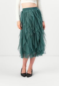Teal tulle skjørt med lagdelte volanger, midt på leggen lengde, elastisk midjebånd, kombinert med svarte spisse hæler på en nøytral bakgrunn.