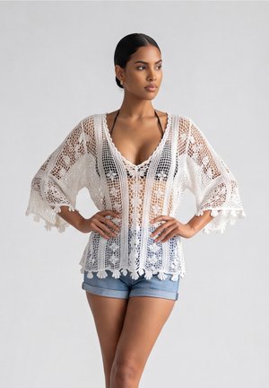 Vrouw draagt een witte kanten gehaakte blouse over een zwart bikinibovenstuk en blauwe spijkershorts, staand met handen in de zij tegen een effen achtergrond.