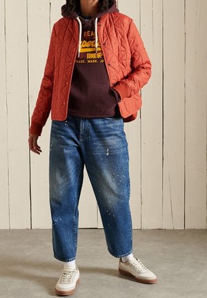 Osoba ubrana w pikowaną kurtkę w kolorze rdzawego pomarańczu, na brązowej bluzie z kapturem z żółtym napisem, niebieskie dżinsy poplamione farbą, białe skarpetki i beżowe buty sportowe.