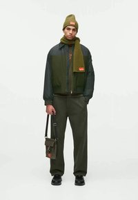 Casaco bomber verde-azeitona com punhos canelados, gorro e cachecol canelados a combinar, calças verde-azeitona escuras e uma pequena bolsa de ombro com um rótulo laranja.