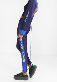 Granatowy krótki top z odkrytymi plecami, zestawiony z niebieskimi legginsami w kratkę z napisem 'WET', uzupełniony czarnymi sneakersami na platformie.